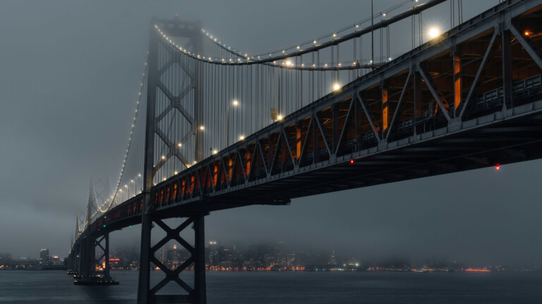 A stunning 4K wallpaper featuring the San Francisco Bay Bridge at dusk. The bridge is beautifully illuminated against a backdrop of the vibrant evening sky, capturing the city's iconic skyline. Ideal for setting as your desktop or mobile wallpaper.
