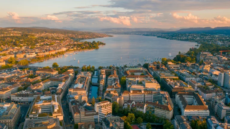 A mesmerizing 4K wallpaper captures the enchanting beauty of Zurich at sunset from an aerial perspective. The city's skyline is illuminated by the warm hues of the setting sun, casting a golden glow over the urban landscape. Lights from buildings twinkle as dusk settles in, creating a picturesque scene ideal for adorning your desktop or mobile screen.