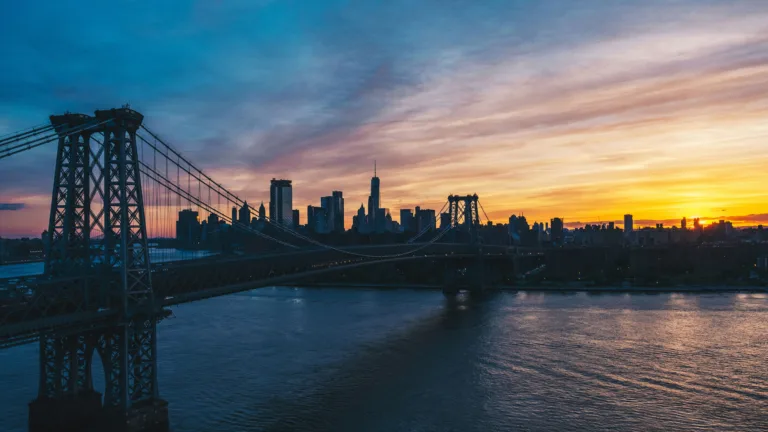 A stunning 4K wallpaper featuring the Williamsburg Bridge at sunset, with the New York skyline silhouetted against a vibrant evening sky. The warm hues of the sunset reflect off the water, creating a picturesque and serene scene perfect for your desktop or mobile background.