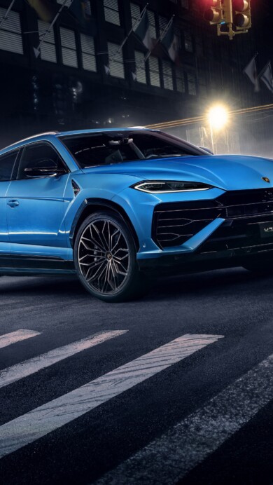 A captivating 4K wallpaper featuring a Lamborghini Urus cruising through a vibrant city street at dusk. The sleek design of the luxury SUV is highlighted against the urban backdrop, showcasing its power and elegance.