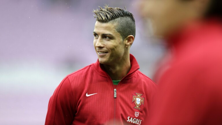 A vibrant 4K wallpaper showcases Cristiano Ronaldo in a red Portugal national team training jacket, smiling softly as he looks off to the right. Fine rain droplets are visible on his spiky hair and the subtly blurred purple-grey background, adding a calm, atmospheric depth to the portrait.