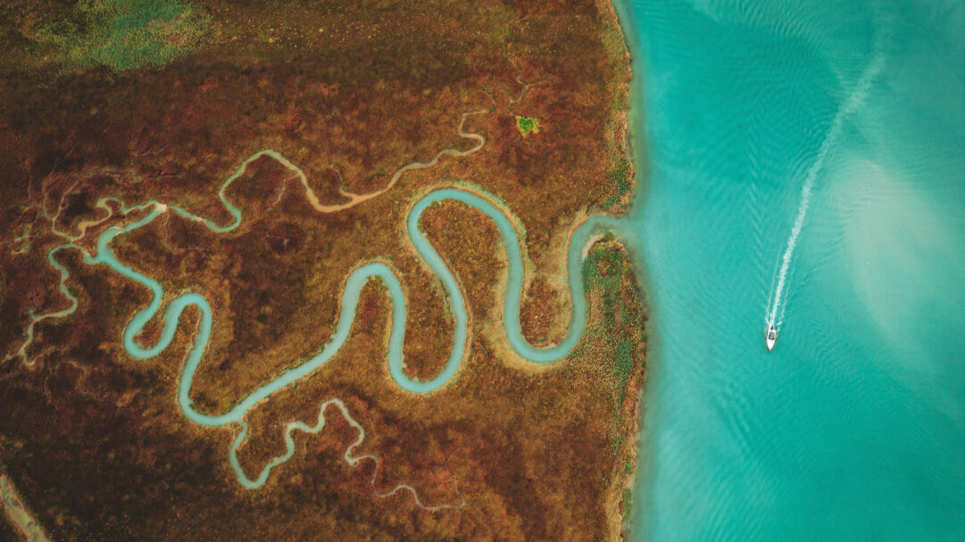A captivating 4K wallpaper unveils an aerial view of the Venetian Lagoon's intricate turquoise delta waterways winding through a rust-toned marshland. The vibrant turquoise of the winding delta channels and the broad lagoon contrasts dramatically with the rich, rust-colored marsh, where a lone white boat leaves a crisp wake.