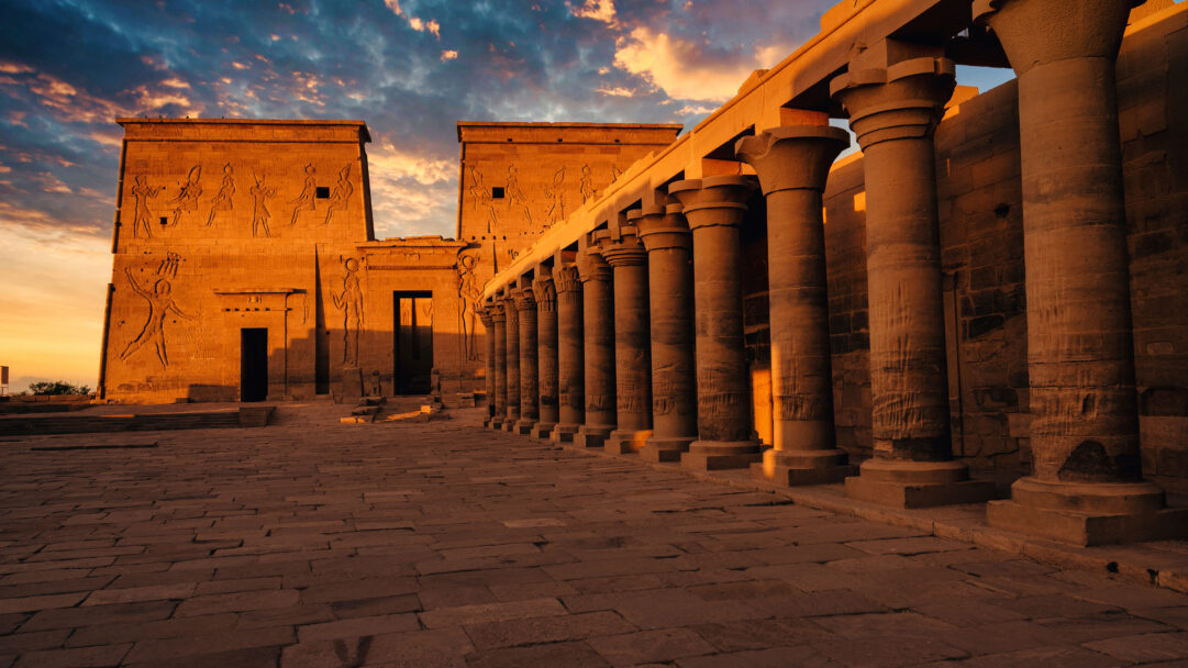 A captivating 4K wallpaper showcases the ancient Philae Temple in Aswan, Egypt, bathed in the warm glow of sunset. Golden sunlight illuminates its grand columns and intricately carved walls, casting long shadows across the ancient stone and creating a serene, majestic atmosphere.