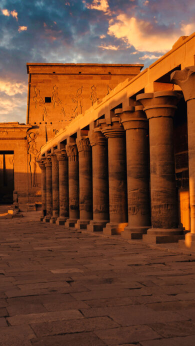 A captivating 4K wallpaper showcases the ancient Philae Temple in Aswan, Egypt, bathed in the warm glow of sunset. Golden sunlight illuminates its grand columns and intricately carved walls, casting long shadows across the ancient stone and creating a serene, majestic atmosphere.