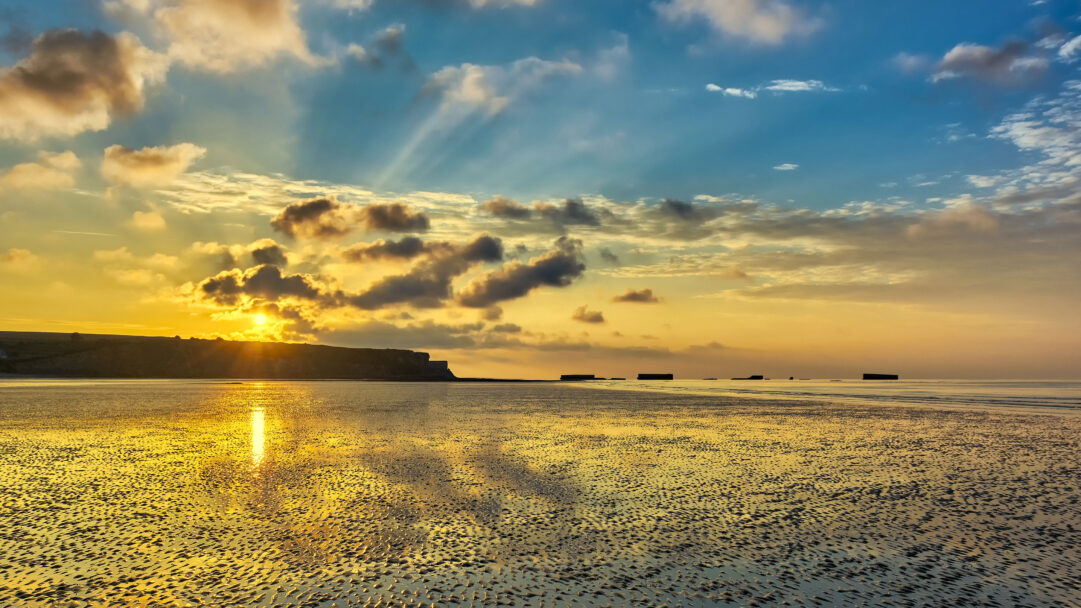 A breathtaking 4K wallpaper capturing the Arromanches sunset over golden sands, featuring the distant, iconic Mulberry Harbour remains. The low sun bathes the textured wet sand in a vibrant golden glow, mirroring the radiant sky and emphasizing the tranquil yet historically significant seascape.