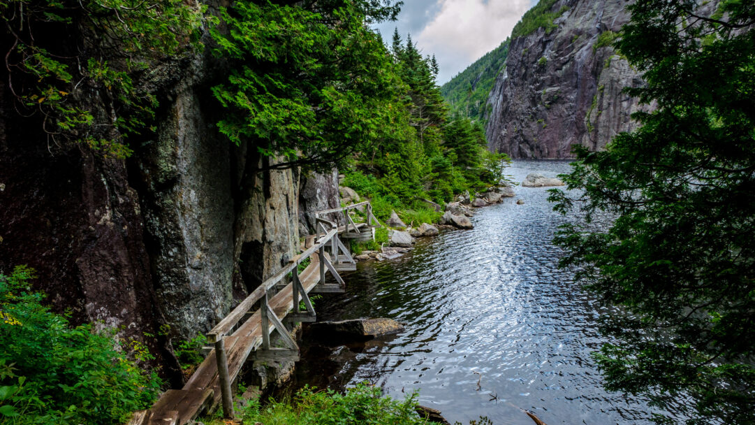 A scenic 4K wallpaper showcasing the Avalanche Lake Trail with its wooden path winding along a steep cliff face in the Adirondack High Peaks. The shimmering dark waters of Avalanche Lake reflect the towering rocky cliffs and dense green forest, creating an immersive and tranquil outdoor escape.