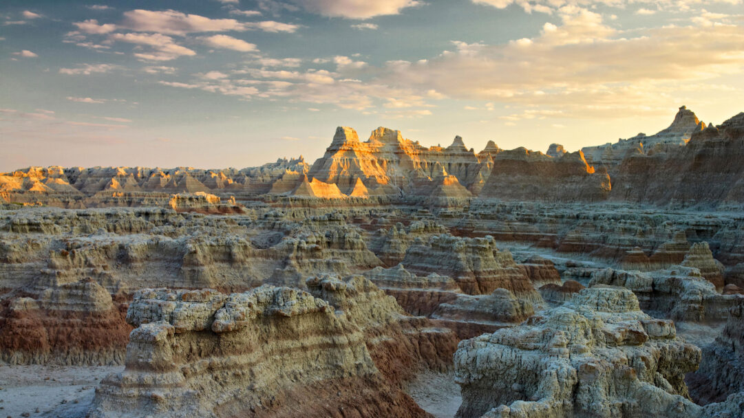 A breathtaking 4K wallpaper showcasing the rugged, eroded landscape of Badlands National Park in South Dakota at sunset. Golden light illuminates the intricate peaks and buttes, contrasting sharply with the deep shadows and revealing the rich, stratified colors of the terrain.