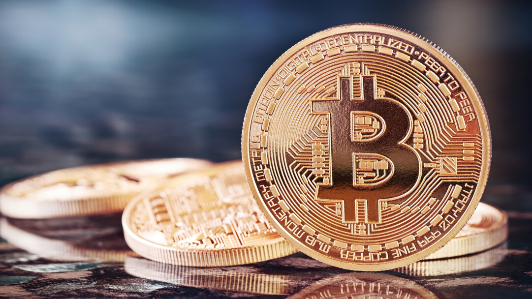 A brilliant 4K wallpaper capturing a detailed gold Bitcoin coin standing prominently on a reflective dark surface, with other blurred coins nestled behind it. The coin's intricate circuitry patterns and warm metallic glow are beautifully highlighted by subtle reflections and a serene, blurred blue backdrop, evoking a sense of digital elegance. A shiny, golden Bitcoin cryptocurrency coin stands upright, prominently displaying its 'DECENTRALIZED DIGITAL PEER TO PEER' text and intricate circuitry patterns. Other blurred gold coins lie on a reflective surface behind it, set against a dark, soft background.