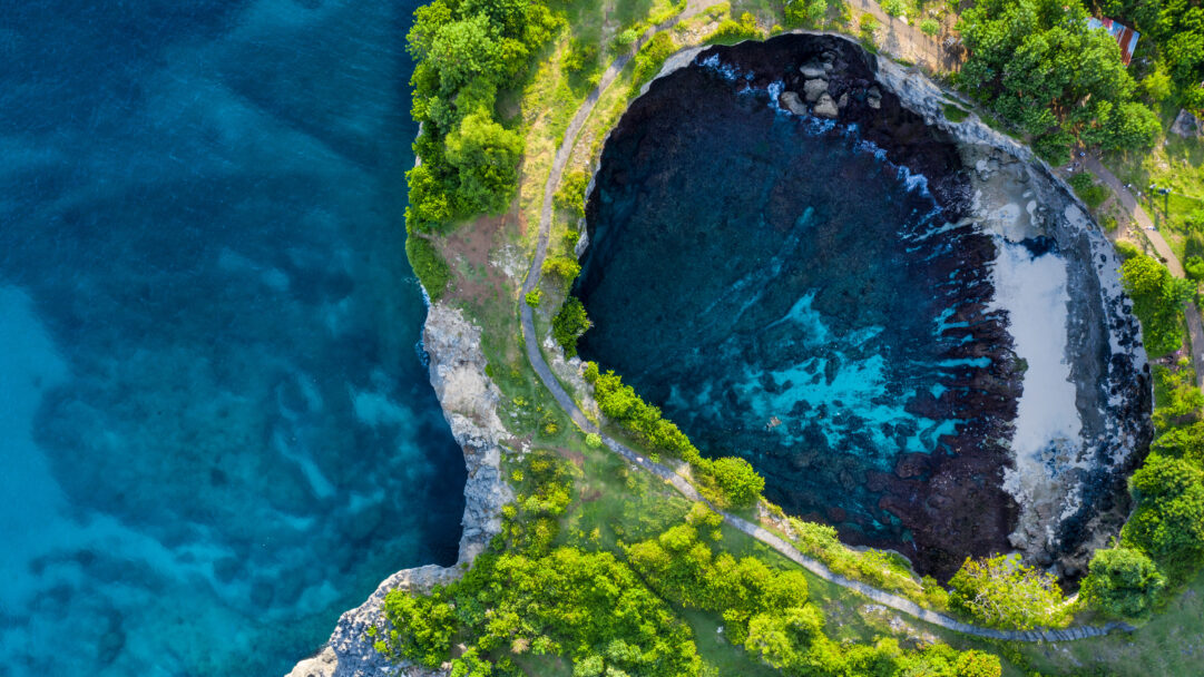 A captivating 4K wallpaper offering an aerial view of the iconic Broken Beach on Nusa Penida, Bali, Indonesia, where a pristine turquoise lagoon is encircled by verdant cliffs. The stark contrast between the vibrant green foliage, the deep blue open ocean, and the clear shallow waters within the natural circular cove creates an inviting, serene tropical escape.