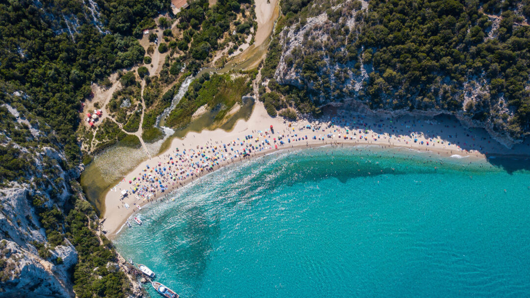 A stunning 4K wallpaper showcasing Cala Luna Beach, Sardinia's renowned turquoise paradise, with its crescent of golden sand bordered by lush green cliffs and a winding river. The spectacularly clear, brilliant turquoise waters are alive with sunlight, inviting numerous bathers into their sparkling depths.