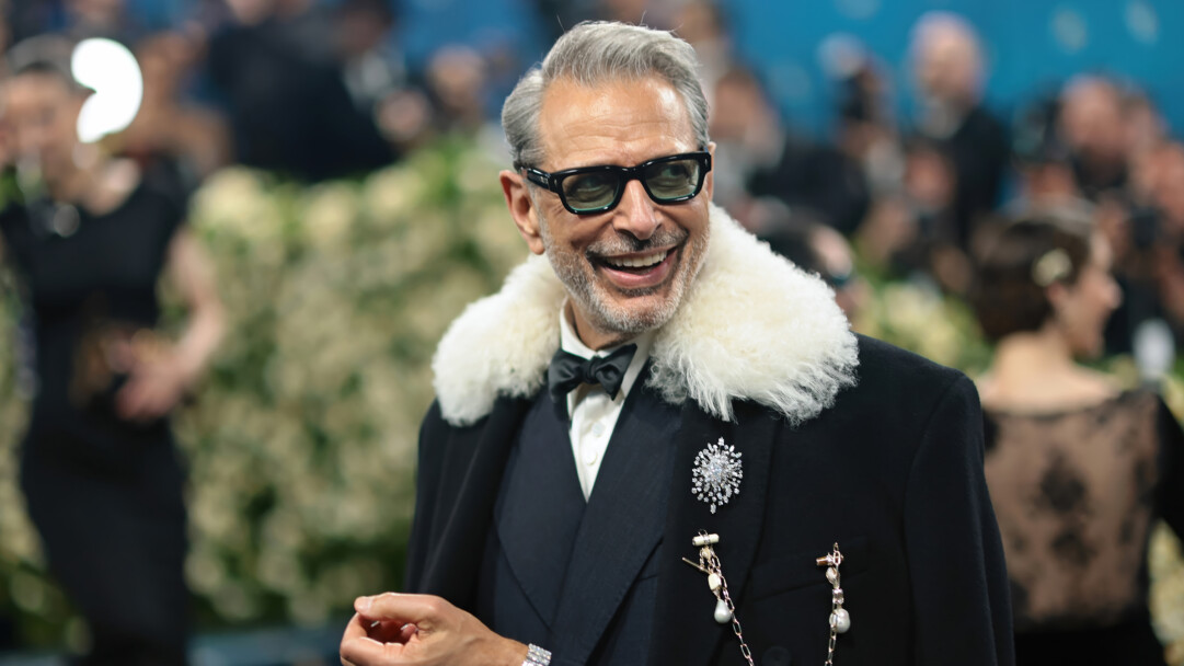 An iconic 4K wallpaper features a charming Jeff Goldblum smiling widely at the Met Gala, dressed in a distinctive black suit with a prominent white fluffy collar. His dark sunglasses and sparkling brooches enhance his eccentric elegance, reflecting a joyful and approachable mood.