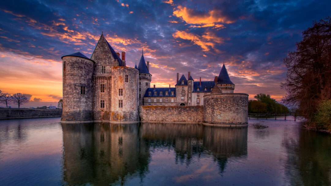 A captivating 4K wallpaper presents the majestic Château de Sully-sur-Loire in Centre-Val de Loire, grandly positioned within its reflective moat. The dramatic sunset sky bursts with fiery orange and deep indigo clouds, casting an enchanting glow over the ancient stone and mirroring brilliantly in the still waters below.