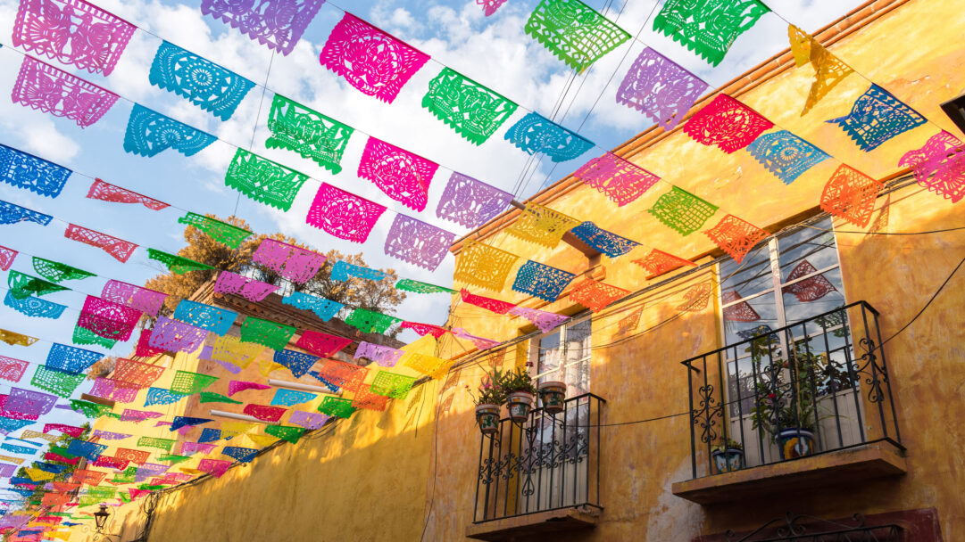 A vibrant 4K wallpaper showcasing colorful papel picado, intricately cut paper flags, strung vibrantly across a charming street in San Miguel de Allende. These festive banners cast dynamic shadows against a warm golden-hued building, creating a cheerful and lively atmosphere under the bright sky.