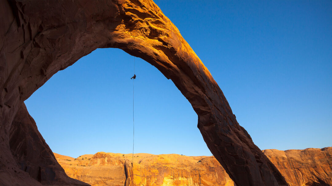 An adventurous 4K wallpaper featuring a lone figure rappelling from the iconic Corona Arch, set against a vast desert sky. Golden hour sunlight dramatically illuminates the massive sandstone arch and distant desert cliffs, casting deep shadows that accentuate the grandeur of this outdoor adventure.
