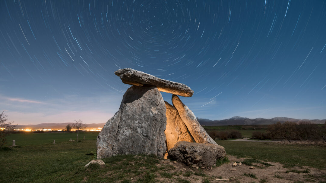 A captivating 4K wallpaper captures the ancient Dolmen of Sorginetxe standing majestically in a grassy field under the night sky of the Basque Country, Álava. Long, arcing star trails swirl across the deep blue expanse, beautifully contrasting with the warm, golden light emanating from within the ancient stone structure.
