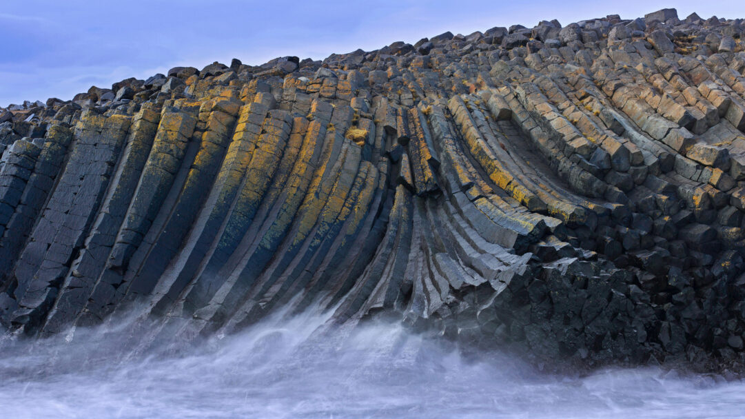 A majestic 4K wallpaper showcasing dramatic basalt columns rising from the turbulent North Atlantic at Kálfshamarsvík, Iceland. The powerful hexagonal rock formations, partially covered in golden lichen, meet the swirling, long-exposure ocean mist, creating a scene of raw, untamed natural grandeur.