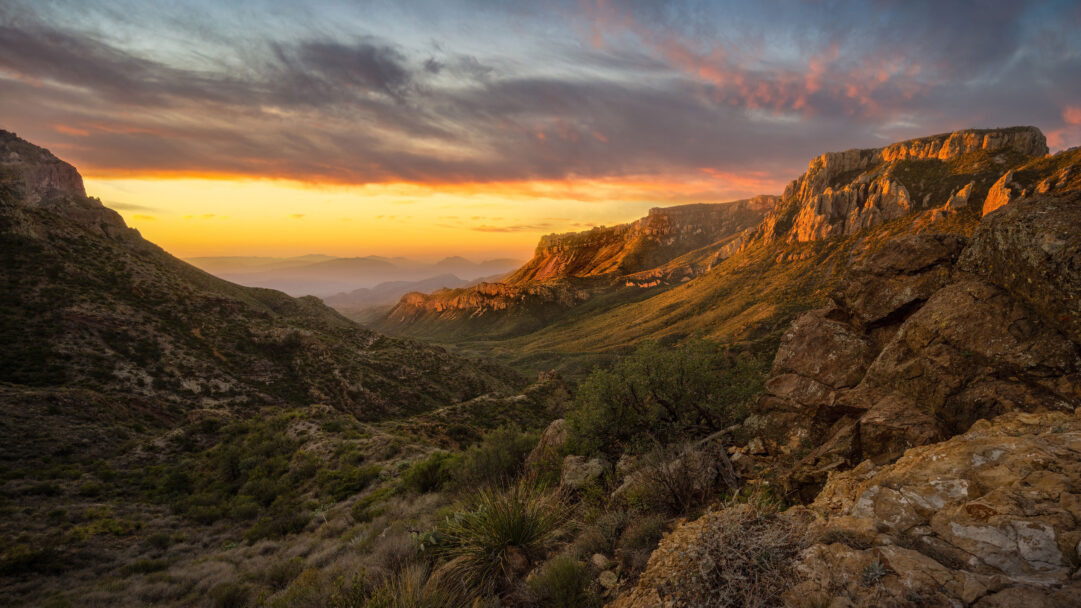 A vibrant 4K wallpaper showcases the dramatic landscape of Big Bend National Park, with the Chisos Mountains bathed in the intense light of a setting sun. The golden sunset vividly illuminates the rugged peaks and deep valleys, creating a breathtaking interplay of light and shadow that highlights the desert terrain and a colorful, streaked sky.