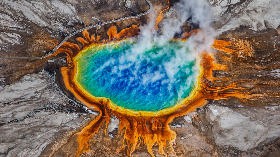 A magnificent 4K wallpaper showcases Yellowstone's Grand Prismatic Spring from an aerial perspective, revealing its massive, vividly colored thermal pool and surrounding mineral terraces. Steaming with ethereal mists, the spring's deep blue center transitions through brilliant teal, green, and fiery orange rings, creating a breathtaking natural tapestry of vibrant hues.