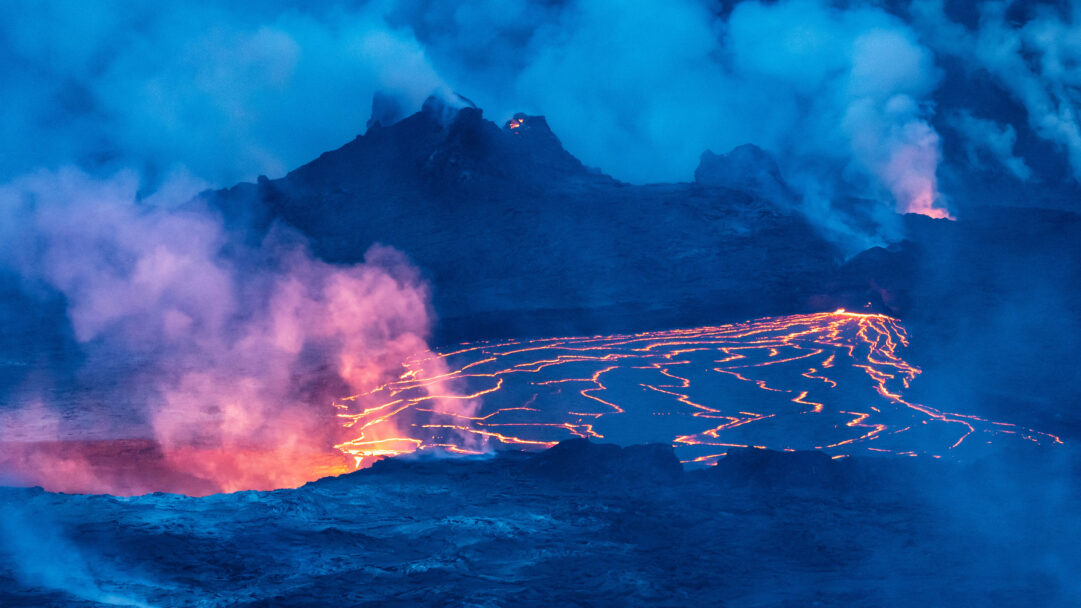 A mesmerizing 4K wallpaper of Halema'uma'u Crater's lava lake at night, showcasing an active eruption within the vast, dark caldera. Vibrant streams of fiery orange lava illuminate the scene, contrasting sharply with the deep blue volcanic landscape and plumes of glowing, reddish-pink steam rising into the night sky.