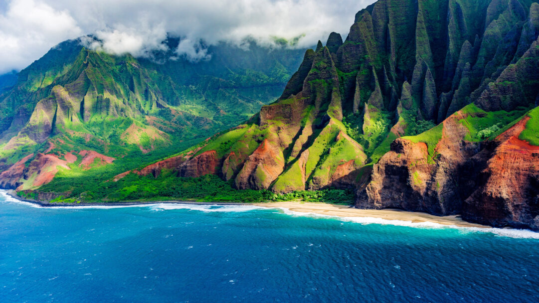 A breathtaking 4K wallpaper captures Hawaii’s majestic Na Pali Cliffs rising dramatically above Kalalau Beach and its vibrant turquoise waters. Sunlight illuminates the richly textured, emerald and ochre cliffs, contrasting beautifully with the deep azure ocean, creating an awe-inspiring natural vista.