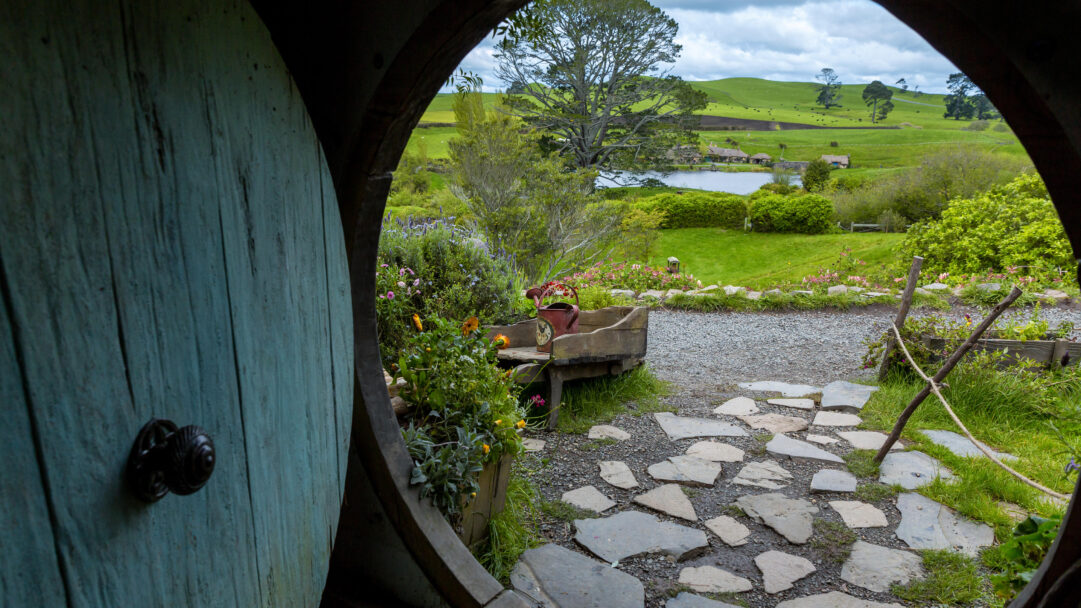 A magical 4K wallpaper offers a picturesque landscape view from inside a Hobbit-hole door at the Hobbiton Movie Set. The vibrant green of the rolling hills, a tranquil lake, and distant Hobbit-holes are beautifully framed by the rustic turquoise door on the left, evoking a sense of peaceful wonder.