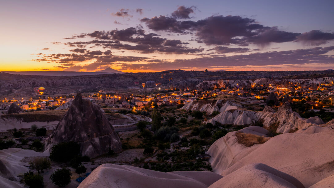 A magical 4K wallpaper of hot air balloons gracefully ascending over the illuminated town of Goreme in Cappadocia. The dramatic sunset paints the sky with hues of orange and purple, casting a warm glow over the distinctive fairy chimneys and the twinkling city lights below.