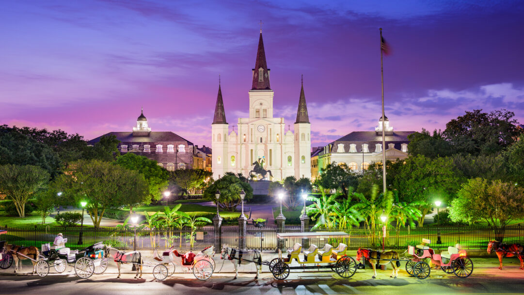 An enchanting 4K wallpaper of St. Louis Cathedral majestically dominating Jackson Square in New Orleans, Louisiana, during a tranquil evening, with several horse-drawn carriages lining the street in the foreground. The scene is cast under a breathtaking gradient twilight sky of deep purples and pinks, illuminated by the bright white facade of the cathedral and the warm glows from the square's gas lamps.
