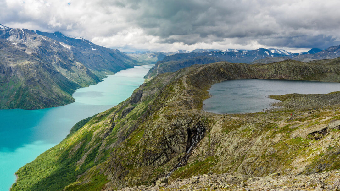 A dramatic 4K wallpaper reveals the iconic Besseggen Ridge in Jotunheimen National Park, Norway, featuring two contrasting mountain lakes amidst a vast mountainous landscape. The vivid turquoise of Lake Gjende sharply contrasts with the deep blue of Lake Bessvatnet, highlighted by the rugged, moss-covered ridge under a dramatic, cloudy sky.