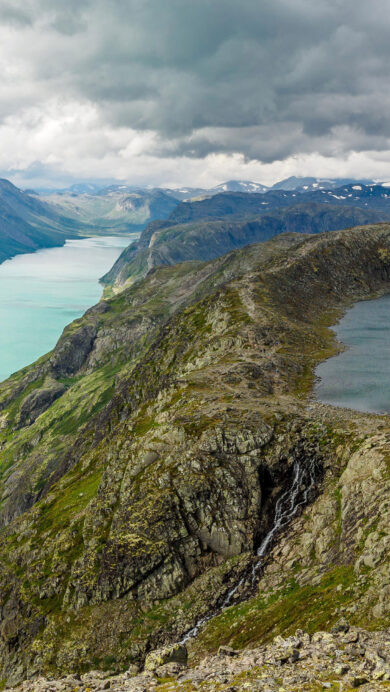 A dramatic 4K wallpaper reveals the iconic Besseggen Ridge in Jotunheimen National Park, Norway, featuring two contrasting mountain lakes amidst a vast mountainous landscape. The vivid turquoise of Lake Gjende sharply contrasts with the deep blue of Lake Bessvatnet, highlighted by the rugged, moss-covered ridge under a dramatic, cloudy sky.