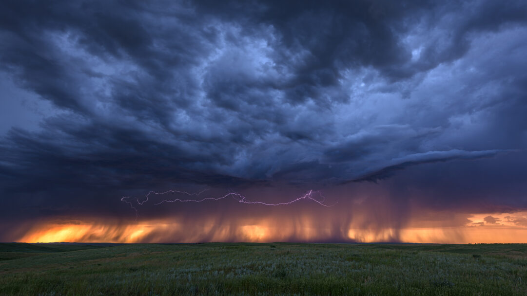 An electrifying 4K wallpaper featuring a powerful lightning storm raging over the expansive green fields of Bowman Field at sunset. Jagged lightning bolts horizontally tear through dark, heavy storm clouds, brightly illuminating dense rain as the setting sun casts a vibrant orange and purple glow across the distant horizon.