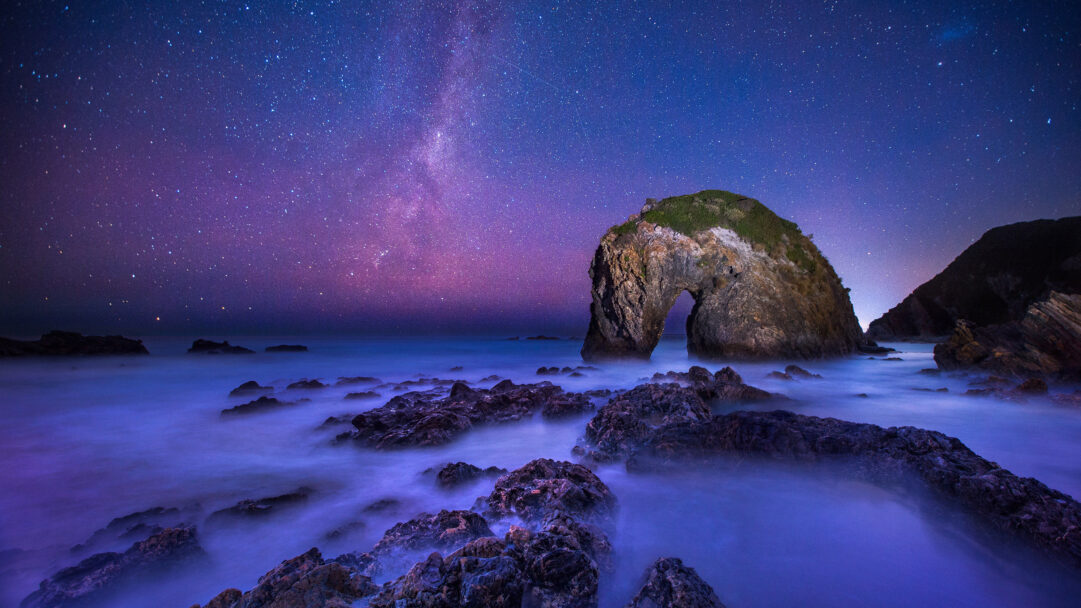 A magical 4K wallpaper of the Milky Way sweeping across a vibrant night sky above the distinct Horse Head Rock on a rugged coastline. The long-exposure capture highlights the soft, misty blue waters swirling around dark rock formations, creating a breathtaking blend of celestial and earthly beauty.