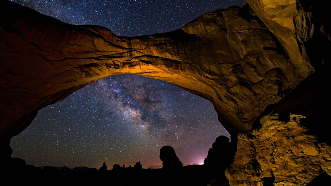 A celestial 4K wallpaper showcases the luminous band of the Milky Way across the vast night sky, perfectly framed by the colossal Double Arch in Arches National Park. The warm, earthy tones of the arch, lit from below, dramatically contrast with the infinite speckled expanse of stars and galactic dust, creating an awe-inspiring vision of the cosmos.