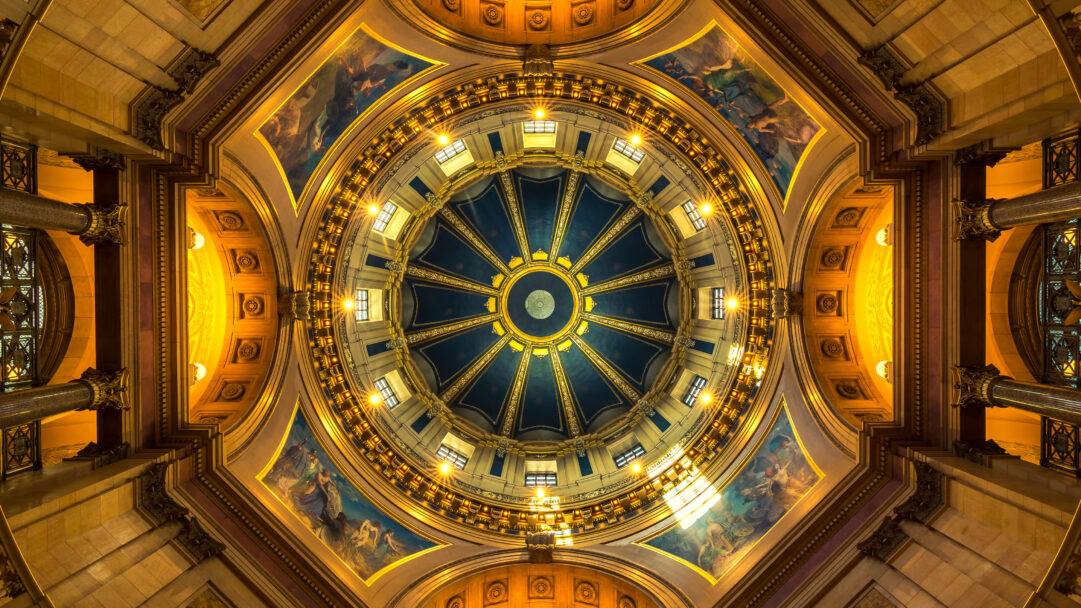 An opulent 4K wallpaper capturing the intricate interior of the Minnesota State Capitol Dome in St Paul, viewed from directly below. Warm golden light bathes the ornate architectural details and vibrant painted murals, radiating outwards from the deep blue central oculus with bright starbursts of light.