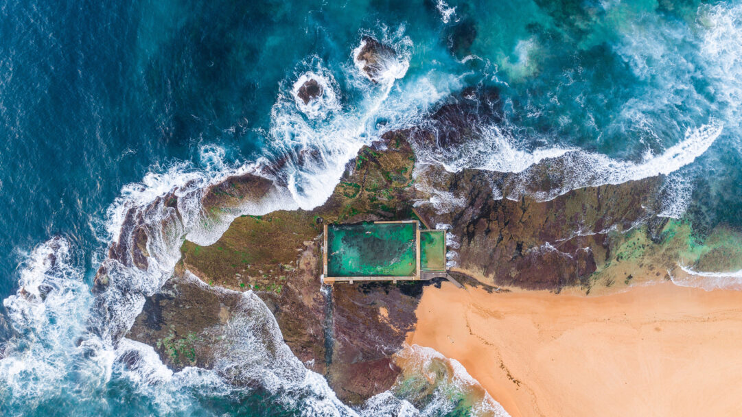 A breathtaking 4K wallpaper presenting an aerial view of the Mona Vale Rockpool nestled against Sydney Beach. The dynamic interplay of deep blue ocean waves, frothy white surf, and a rugged, algae-covered rock shelf creates a dramatic border for the serene, emerald green rockpool and the soft golden beach.