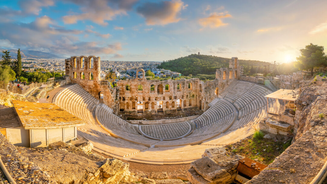 An awe-inspiring 4K wallpaper captures the ancient Odeon of Herodes Atticus, nestled on the slopes of the Acropolis in Athens, overlooking the sprawling city. The warm, golden light of the sunset bathes the historic stone tiers and arched stage, creating a magnificent glow that highlights its ancient grandeur.
