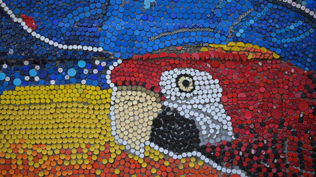 A vibrant 4K wallpaper showcasing Oscar Olivares' Guatire bottle cap parrot, a large-scale mosaic art piece composed entirely of colorful recycled plastic caps. The parrot's prominent red head and bright yellow body are distinctively rendered, with its detailed eye formed by white, black, and yellow caps, creating a striking and textured visual.
