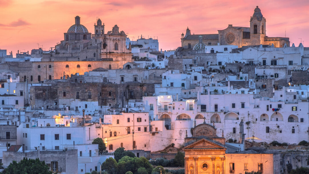 A captivating 4K wallpaper presents the historic city of Ostuni, Apulia, Italy, a dense hillside cityscape dominated by white-washed buildings and ancient architectural structures. The scene is beautifully illuminated by the warm, golden-pink hues of dusk, casting a soft glow over the upper buildings and accentuating the city's unique charm and serene atmosphere.