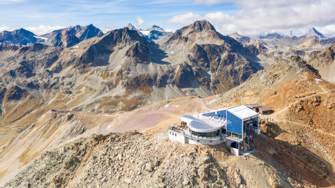 A breathtaking 4K wallpaper showcasing the modern Piz Nair Mountain Station perched high on a rugged peak, surrounded by a vast panoramic expanse of the Swiss Alps. Its sleek, glass-fronted design, featuring prominent blue solar panels, stands in stark, awe-inspiring contrast to the raw, barren landscape and distant snow-dusted mountains under a clear blue sky.