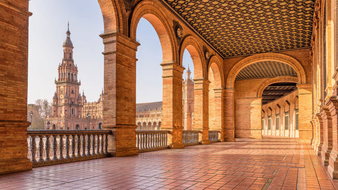 A sun-drenched 4K wallpaper capturing the grand colonnade architecture of Plaza de España in Seville, Spain, where a series of arches frames a view of a distant, ornate tower. The warm golden light beautifully illuminates the intricate brickwork and the ornate geometric ceiling, creating a captivating interplay of shadows on the tiled floor.