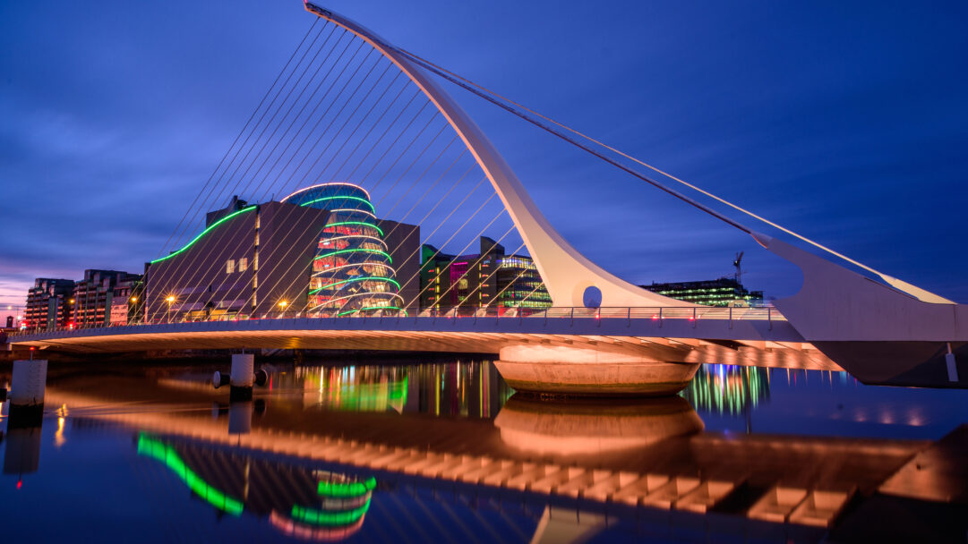 A breathtaking 4K wallpaper showcasing the Samuel Beckett Bridge gracefully arching over the River Liffey in Dublin, surrounded by the glow of the night cityscape. Its distinctive harp-like structure is vibrantly illuminated, casting stunning, colorful reflections of itself and the modern buildings onto the tranquil water.