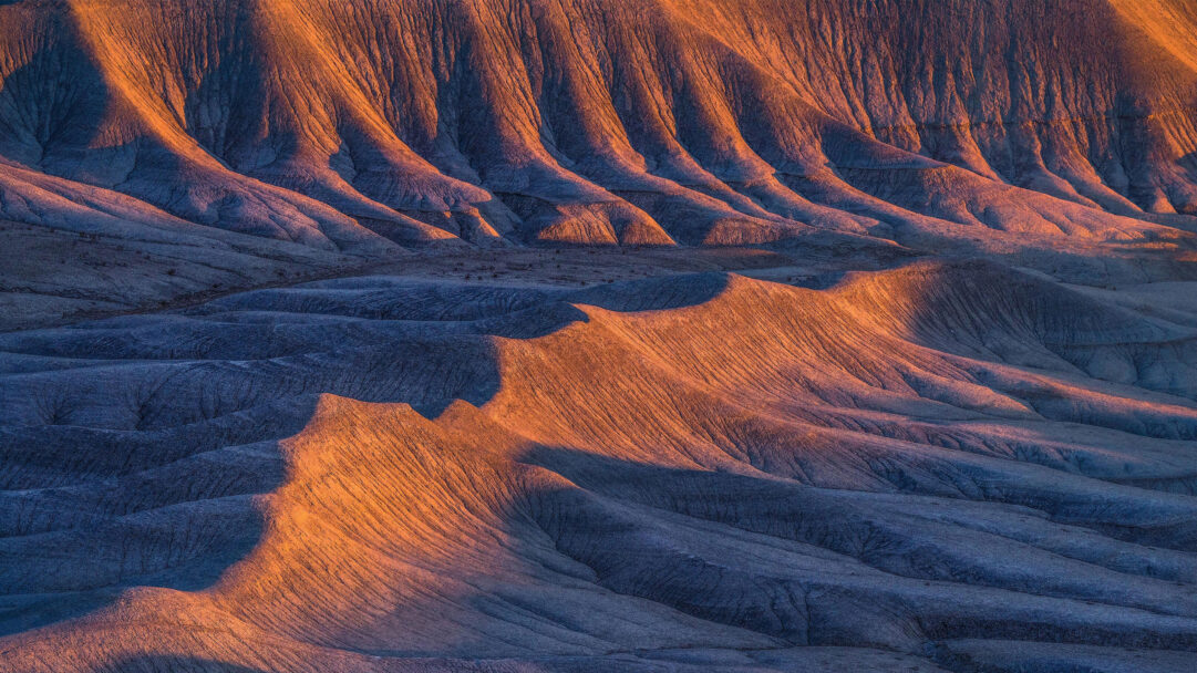 A captivating 4K wallpaper showcases dramatic sandstone formations in the Caineville Badlands, illuminated by the warm glow of sunset. Golden light highlights the intricate, ridged textures of the peaks, creating striking contrasts with the cool, deep blue shadows filling the valleys, defining the landscape's rugged beauty.