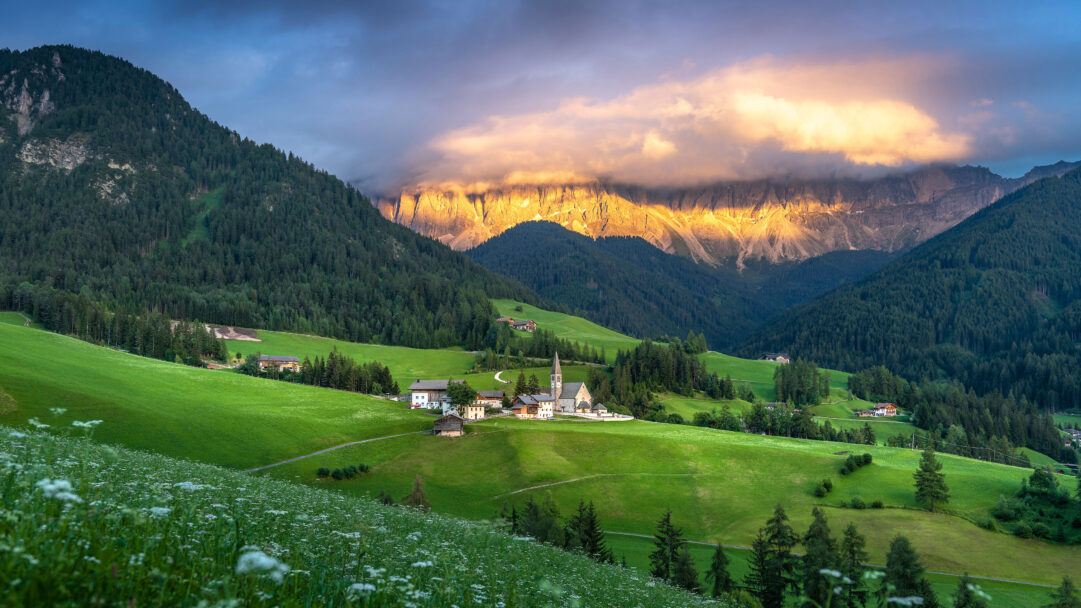 A breathtaking 4K wallpaper captures the picturesque Santa Maddalena village, complete with its iconic church spire, nestled serenely within a verdant valley in the Dolomites. The dramatic golden hour light brilliantly illuminates the towering Dolomite peaks in the background, creating a majestic contrast with the cooler, shadowed valley and enhancing the tranquil atmosphere.