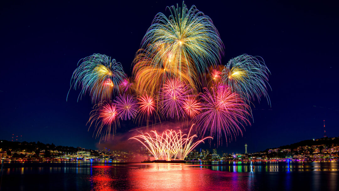 A spectacular 4K wallpaper captures a vibrant fireworks display over Seattle's Lake Union at night, framed by the illuminated city skyline. Brilliant bursts of red, magenta, gold, and teal fireworks fill the dark sky and reflect dynamically on the calm water, creating a festive and dazzling urban spectacle.