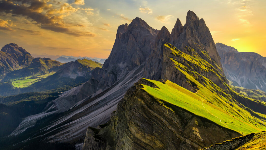 An epic 4K wallpaper showcases the iconic Seceda Peak in the Dolomites, magnificently illuminated during a vibrant sunset. The golden light brilliantly highlights the jagged grey ridges and the intensely green, sun-drenched slopes, creating a powerful contrast and a sense of awe.