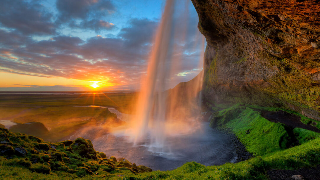 An immersive 4K wallpaper capturing Seljalandsfoss Waterfall in Iceland, as it cascades powerfully from a rugged, moss-covered cliff face into a shadowy basin below. The vibrant sunset paints the sky with fiery orange and soft blues, while its golden light streams through the waterfall's mist, creating an ethereal glow that highlights the surrounding lush green landscape.