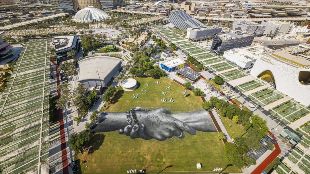 A panoramic 4K wallpaper showcases Saype's 'beyond Walls' land art, featuring two giant clasped hands painted on a lush green lawn at the sprawling Expo 2020 Dubai site. The detailed, monochrome hands stand out dramatically against the vibrant grass, conveying a powerful message of global unity and human connection.