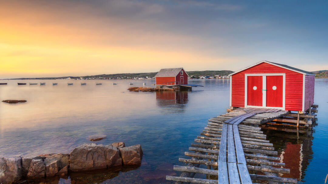 A mesmerizing 4K wallpaper capturing the sunset over a tilting fishing village on Fogo Island, where vibrant red fishing sheds stand on weathered stilts in the calm bay. The warm, diffused light of the setting sun bathes the scene in hues of gold and blue, reflecting softly on the water and highlighting the rustic charm of these unique structures.