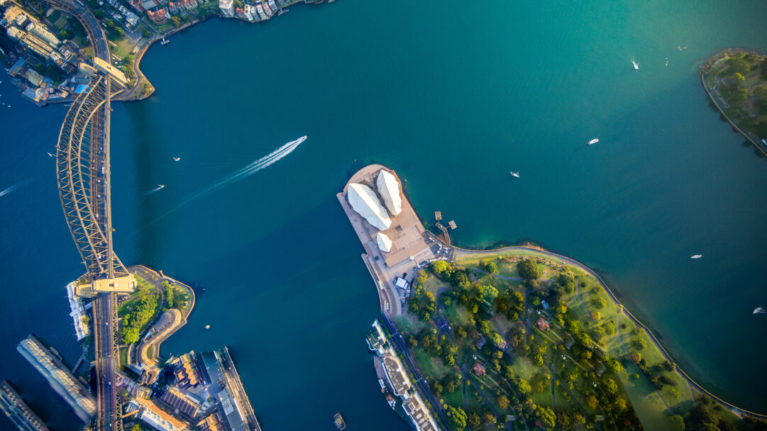 A breathtaking 4K wallpaper capturing an expansive aerial view of Sydney Harbour, prominently featuring the iconic Sydney Harbour Bridge spanning the water and the distinctive Sydney Opera House on the shore. The deep teal waters of the harbour are beautifully illuminated by golden sunlight, reflecting the vibrant life of boats moving across the surface, creating a dynamic and serene cityscape.