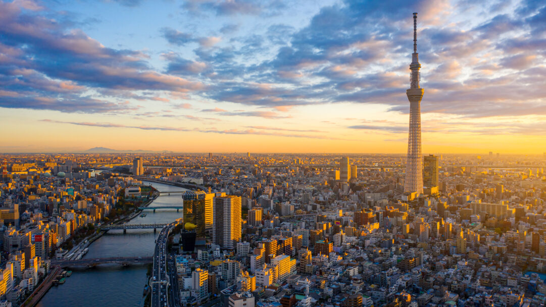 A breathtaking 4K wallpaper captures an expansive aerial view of the Tokyo cityscape at sunrise, with the iconic Tokyo Skytree dominating the right side. Golden sunlight illuminates the myriad buildings and a winding river, creating a warm glow that contrasts beautifully with the soft blue and pink clouds in the morning sky.