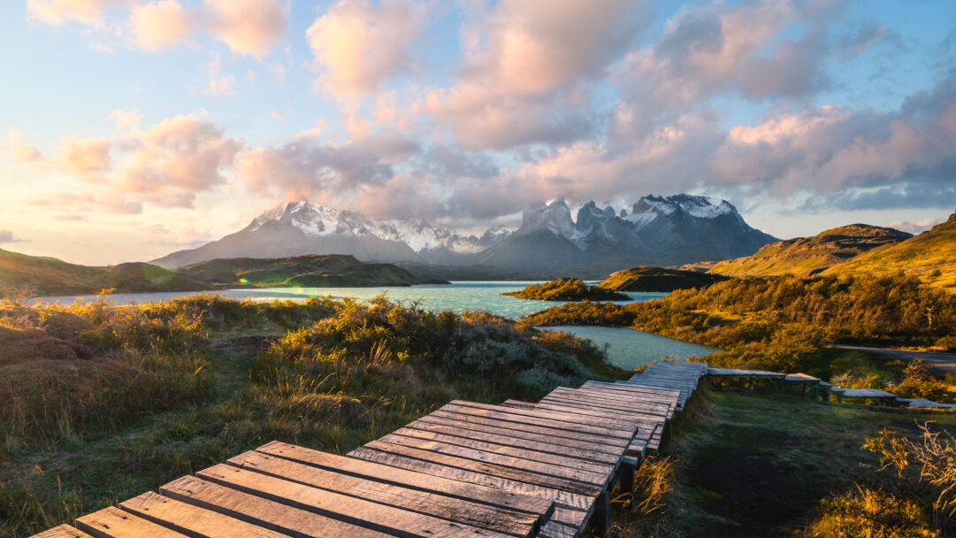 A breathtaking 4K wallpaper captures a serene sunset landscape in Torres del Paine National Park, featuring a tranquil lake, majestic snow-capped mountains, and a curving wooden boardwalk. The golden hour light beautifully illuminates the foreground boardwalk and surrounding grasses, creating a warm contrast with the cool turquoise water and the dramatic, cloud-draped peaks, evoking a sense of peaceful grandeur.